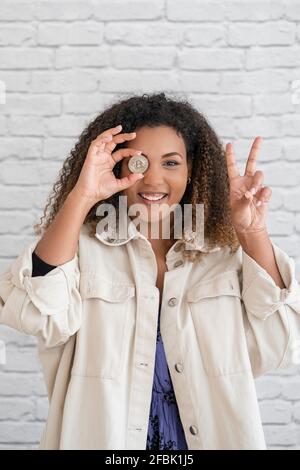 Lächelnde junge Frau, die ein Friedenszeichen zeigt, während sie vor der Wand das Auge mit Bitcoin bedeckt Stockfoto