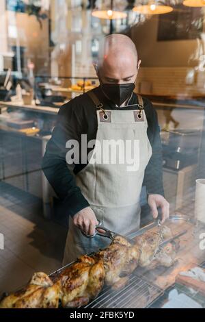 Der Chefkoch schneidet im Restaurant gebratenes Hähnchen, das durch das Glas gesehen wird Küche Stockfoto