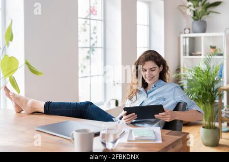 Lächelnde Unternehmerin, die mit einem digitalen Tablet sitzt Füße hoch am Schreibtisch Stockfoto