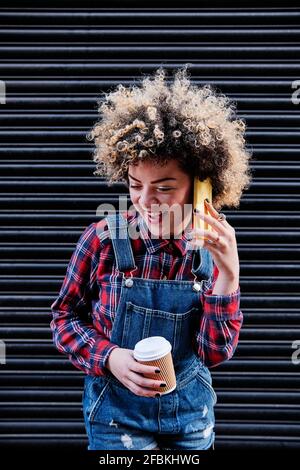 Lockig haarige Frau mit Einweg-Kaffeetasse lachend während des Sprechens Auf dem Mobiltelefon Stockfoto