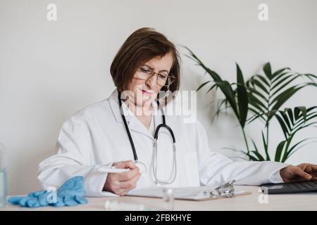 Ältere Ärztin liest den Bericht, während sie an der Rezeption sitzt Klinik Stockfoto