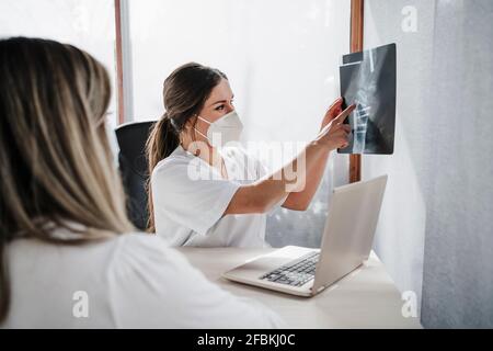 Ärztin trägt eine schützende Gesichtsmaske, die dem Patienten eine Röntgenaufnahme zeigt Am Schreibtisch in der Klinik Stockfoto