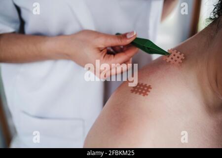 Weibliche Physiotherapeutin klebt mit einer Pinzette ein elastisches Kreuzband an Die Schulter des Patienten in der Klinik Stockfoto