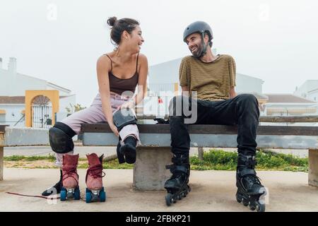 Fröhliche Freundin beim Sitzen auf Freund mit Rollschuhe schauen Auf der Bank Stockfoto