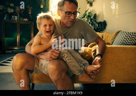 Vater schaut auf den Fuß des weinenden Sohnes, während er auf dem Sofa sitzt Zu Hause Stockfoto