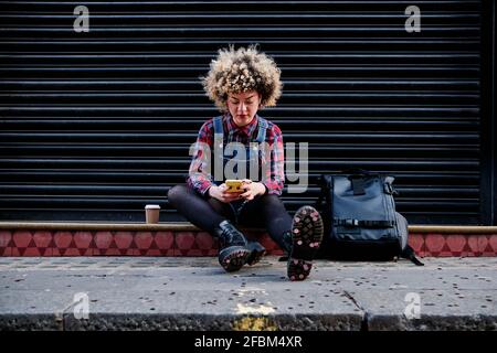 Junge Frau, die Smartphone benutzt, während sie auf dem Fußweg sitzt Stockfoto