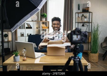 Stilvoller afrikanischer Mann beim Auspacken neuer Sneakers während der Videoaufnahme vor der Kamera. Konzept von Menschen, Bloggen und Werbung. Stockfoto