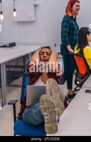Eine nervöse Büroangestellte mit ihren Beinen auf ihrem Schreibtisch und ihrem Laptop in ihrem Schoß, die einen harten Tag hatten. Stockfoto