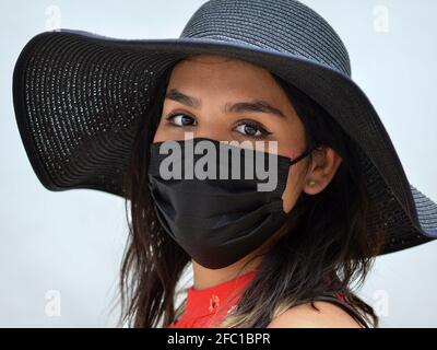 Die junge mexikanische Brünette mit schönen braunen Augen trägt während der Coronavirus-Pandemie einen schwarzen Sonnenhut und eine schwarze Einweg-Gesichtsmaske. Stockfoto