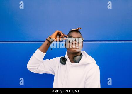 Ein junger afroamerikanischer schwarzer Mann, der ein weißes Sweatshirt, eine Sonnenbrille, einen Kamm und Kopfhörer an einer blauen Wand trägt, ist selbstbewusst Stockfoto