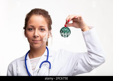 Junge Ärztin oder Krankenschwester hält ein dekoratives Grün hoch Weihnachtskugel in ihrer Hand mit einem freundlichen Lächeln auf Feiern Sie die Weihnachtszeit am Whit Stockfoto