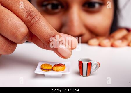 Fun Food Konzept einer Frau, die nach Miniatur-Gebäck greift Und eine Tasse Kaffee auf einem Tisch mit Fokus auf Die Erfrischungen und ihre Finger Stockfoto