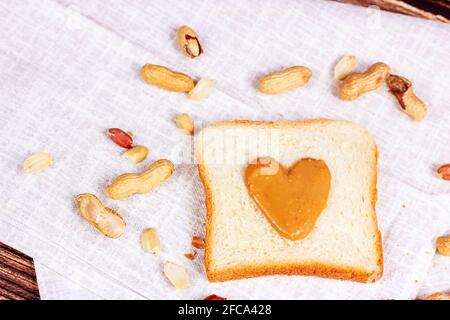 Hausgemachtes Toast zum Frühstück mit frischer, knuspriger, herzförmiger Erdnussbutter und Nüssen auf hellem Textilhintergrund. Stockfoto