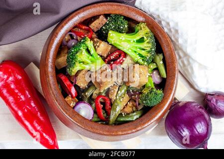 Draufsicht auf hausgemachten vegetarisch gebratenen Tofu mit Gemüse (Brokkoli, Pfeffer, Zwiebeln und grüne Bohnen) in der Holzschüssel auf hellem Küchenhintergrund. Stockfoto