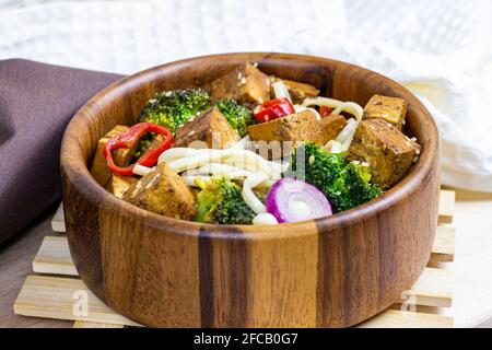 Hausgemachter vegetarischer gebratener Tofu mit udon-Nudeln und Gemüse (Brokkoli, Pfeffer und Zwiebeln) in der Holzschüssel auf hellem Küchenhintergrund. Stockfoto