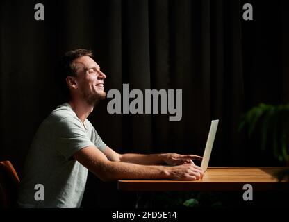 Seitenansicht eines lächelnden Studenten, der am Tisch sitzt und zu Hause am Laptop arbeitet. Konzept des Heimbüros. Stockfoto