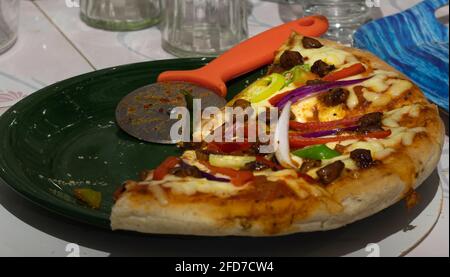 Halbgefressene, in Scheiben geschnittene Pizza auf einem Teller mit Pizzaschneider-Rad in der heimischen Küche, Käse-Hühnchen-Beläge mit Zwiebeln und Paprika und würzig Stockfoto