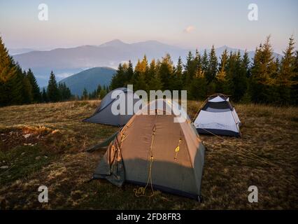 Feriencamp mit drei Zelten auf einer Wiese in Waldnähe. Morgensonne auf Zelt und umliegende Natur. Silhouetten von hohen Bergen auf dem Hintergrund. Stockfoto