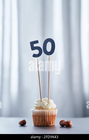 Hausgemachter Vanille-Jubiläums-Cupcake mit cremigem Belag und Nummer 50 fünfzig mit Haselnüssen in der Nähe und hellem Hintergrund. Minimalistisches Geburtstags- oder Jubiläumskonzept. Hochwertiges vertikales Bild Stockfoto