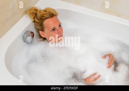 Die blonde Frau mittleren Alters entspannt sich in der Badewanne mit Wasser Wasser und Schaum Stockfoto