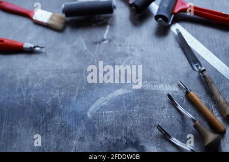 Schneidinstrumente, Tintenroller und andere Werkzeuge für die Linolschnitt-Herstellung mit Kopierraum. Arbeitswerkzeuge in roten und schwarzen Farben auf dunklem Tisch. Konzept der Linolschnitt-Werkzeuge. Hochwertige Fotos Stockfoto