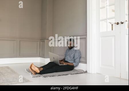 Ein junger afroamerikanischer Student arbeitet zu Hause auf dem Boden sitzend auf einem Laptop. Zu Hause arbeiten oder studieren Stockfoto