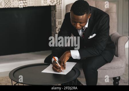 afroamerikanischer Geschäftsmann in einem Anzug macht Notizen auf einem Blatt Papier, während er in seinem Büro in der Nähe des sitzt Kamin Stockfoto