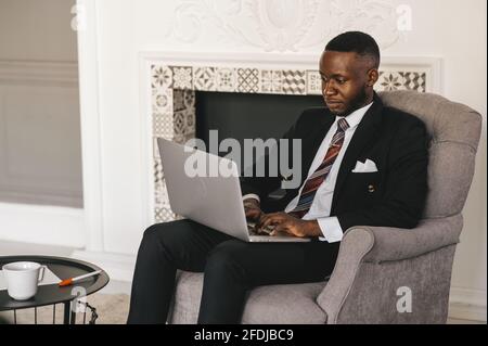 Ein gutaussehender afro-amerikanischer Geschäftsmann im klassischen Anzug verwendet ein Laptop und lächelnd, während Sie im modernen Büro arbeiten Stockfoto