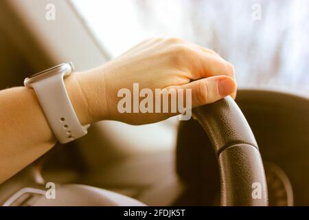 Eine Frauenhand mit einer weißen Uhr hält ein schwarzes Lenkrad aus der Nähe. Verkehr auf einer Straße. Fahrunterricht Konzept. Innenraum des Autos, Speedomete Stockfoto