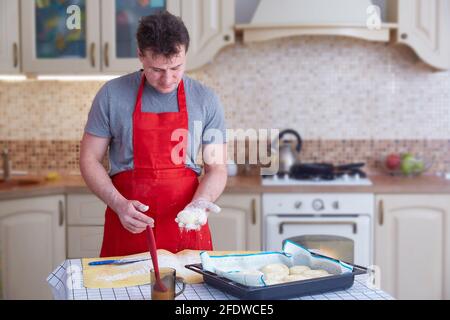 Ein Mann mittleren Alters in einer roten Schürze macht Teigkuchen zu Hause in der Küche. Lifestyle. Speicherplatz kopieren. Stockfoto