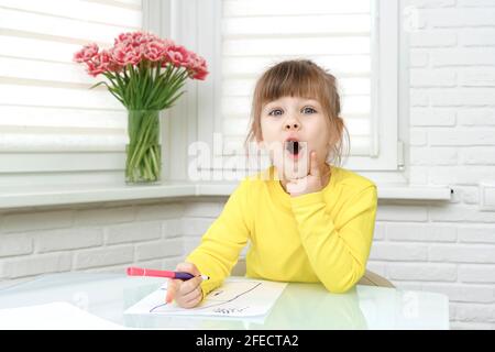 Das kleine Mädchen sitzt an einem Tisch in einem weißen Raum und zeichnet. Stockfoto