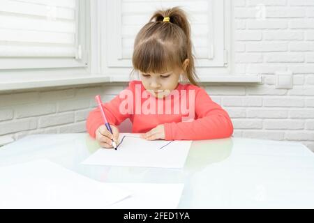 Kleines kaukasisches Mädchen in roten Kleidern sitzt an einem Tisch in einem weißen Raum und zeichnet. Stockfoto