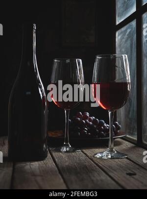 Zwei Gläser Rotwein und eine Flasche neben einem Fenster mit Trauben im Hintergrund auf einem rustikalen Holztisch Stockfoto