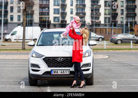 Riga, Lettland - 16. April 2021: Schöne junge Frau mit einem Baby auf den Händen neben einem weißen Auto, die Freude, zusammen zu sein Stockfoto