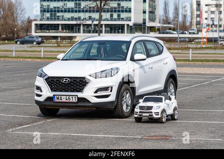 Riga, Lettland - 16. April 2021: Weißer elektrischer Spielzeugwagen für Kinder auf dem Parkplatz neben einem echten Auto Stockfoto