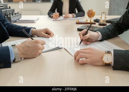 Nahaufnahme von nicht erkennbaren Anwälten in einer Armbanduhr, die sich gegenüber von beiden befindet Andere am Tisch und lesen Vertrag vor der Unterzeichnung bei der Sitzung Stockfoto