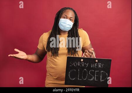 Enttäuschter junger afroamerikanischer Kerl mit Dreadlocks tragen Schutzmaske stehen und halten schwarze Platte mit geschrieben wir sind geschlossen, isoliert auf roter Wand, kleines Unternehmen in der Krise wegen Pandemie Stockfoto