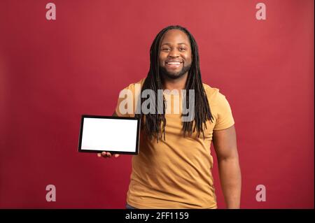Glücklicher afroamerikanischer schwarzer Mann mit Dreadlocks, die isoliert auf rotem Hintergrund stehen, präsentiert neue Anwendung, hält digitale Tablette mit weißem leeren Bildschirm für Werbung, Mockup-Bild Stockfoto
