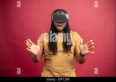 Überrascht verängstigten afroamerikanischen Mixed-Race-Mann mit Dreadlocks und offenen Mund isoliert über rote Wand berühren Luft mit Händen, verbringen Zeit in der virtuellen Realität, spielen Spiele in modernen VR-Headset Stockfoto