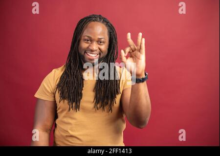 Charismatischer dunkelhäutiger Mann mit langen Dreadlocks, die auf rotem Hintergrund eine OK-Geste zeigen, zeigt ein afrikanischer Mann, dass alles gut ist, keine Sorge Stockfoto