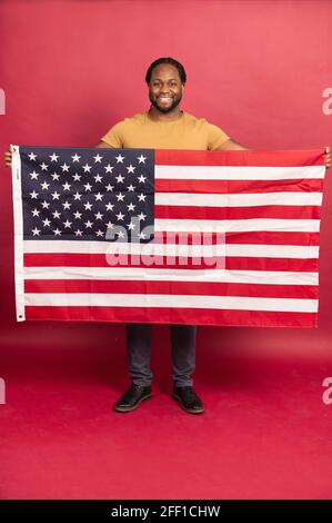 In voller Länge lächelte der schwarz bärtige Mann, der die amerikanische usa-Flagge vor sich hielt, in der Halle auf rotem Hintergrund stand, zeigt ein US-Symbol mit stolzem, vertikalem Schuss, feiert den Unabhängigkeitstag, den 4. Juli Stockfoto