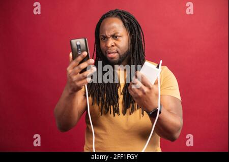 Enttäuschter afroamerikanischer Kerl mit Dreadlocks, die isoliert auf der roten Wand stehen, entladenes Mobiltelefon über USB-Kabel an die Powerbank angeschlossen halten und erfolglos versuchen, das Smartphone aufzuladen Stockfoto