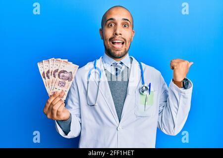 Hispanic erwachsenen Arzt Mann trägt medizinische Uniform hält 500 mexikanische Pesos zeigen den Daumen nach oben auf die Seite und lächeln glücklich mit Öffnen Sie den Mund Stockfoto