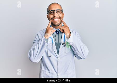 Hispanischer erwachsener Mann mit Ärzteuniform und Stethoskop, lächelnd mit offenem Mund, zeigenden Fingern und einem fröhlichen Lächeln Stockfoto