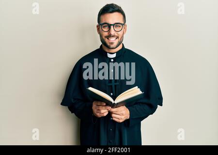 Junger hispanischer Priester, der die bibel hält, lächelt mit einem glücklichen und kühlen Lächeln im Gesicht. Er zeigt Zähne. Stockfoto