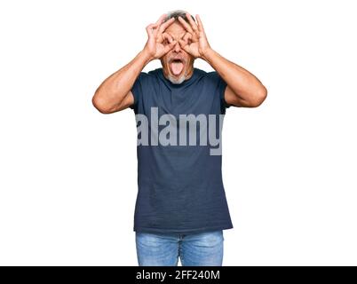 Mittelalter grauhaariger Mann trägt Freizeitkleidung tut ok Geste wie Fernglas aufragende Zunge aus, Augen durch die Finger schauen. Verrückter Ausdruck Stockfoto