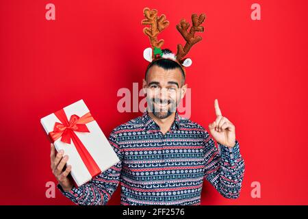 Junger hispanischer Mann trägt Hirsch weihnachtshut mit Geschenk lächelnd mit einer Idee oder Frage Zeigefinger mit glücklichem Gesicht, Nummer eins Stockfoto