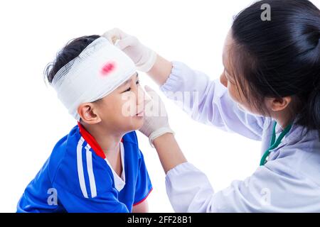 Sportverletzung. Athlet kleiner asiatischer (thailändischer) Junge in blauer Sportkleidung mit Trauma des Kopfes weinend, blutig auf Verband. Doktor macht Verband auf Kopf pati Stockfoto