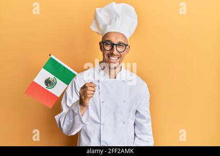 Glatze Mann mit Bart tragen professionelle Koch Schürze hält mexiko Flagge sieht positiv und glücklich stehen und lächeln mit einem Selbstbewusstes Lächeln Stockfoto