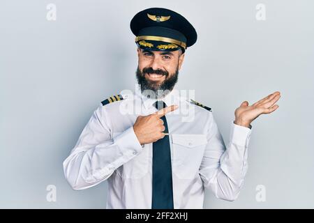 Junger hispanischer Mann in Flugzeugpilotenuniform erstaunt und lächelnd zur Kamera, während er mit der Hand präsentiert und mit dem Finger zeigt. Stockfoto